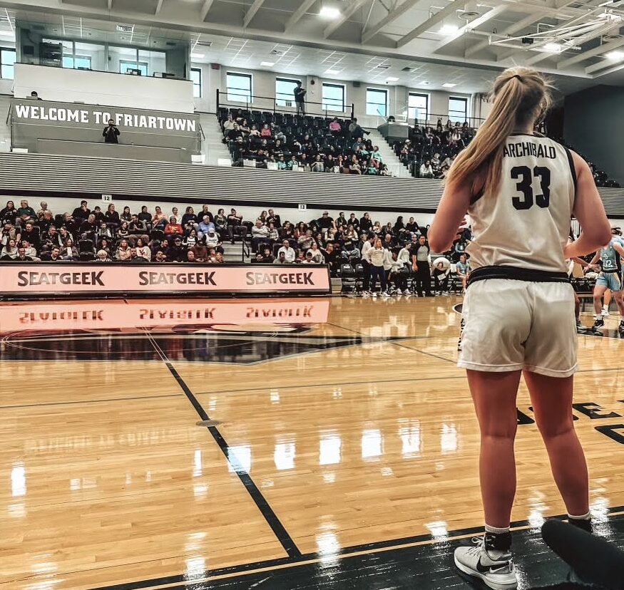 Providence junior forward Emily Archibald inbounds the ball at Providence's home gym, Alumni Hall. She is wearing the Friars' white and black home jersey.